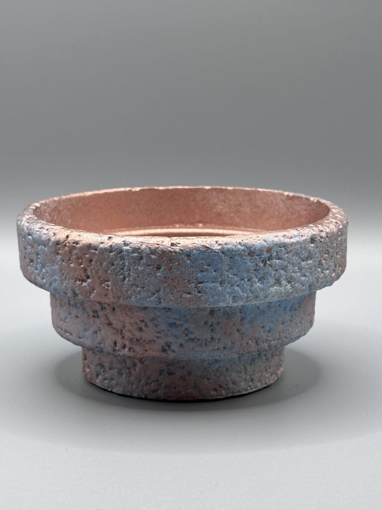A side view of a handcrafted tiered vessel with a heavy, cratered texture and a metallic rose copper and light blue finish against a grey background.