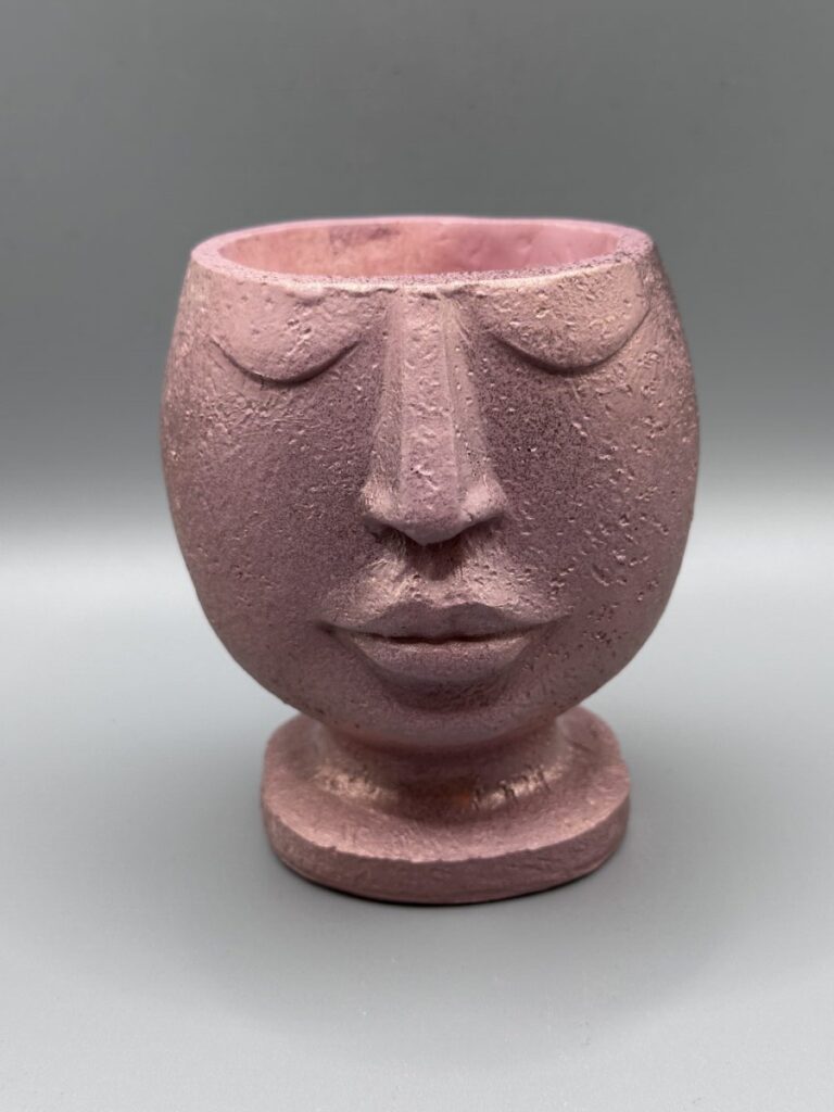 A whimsical feminine face planter with a golden flower in her hair, featuring a peach-toned top and an intricate purple geometric patterned base against a neutral background.