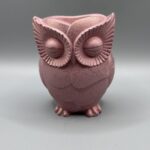 A hand-finished owl planter with closed eyes and detailed feathers, rendered in a metallic rose quartz pink shimmer against a neutral grey background.