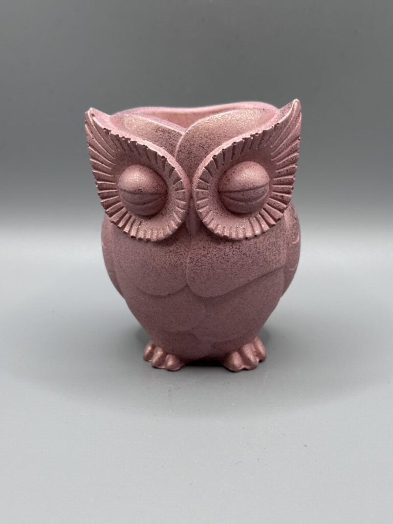 A hand-finished owl planter with closed eyes and detailed feathers, rendered in a metallic rose quartz pink shimmer against a neutral grey background.