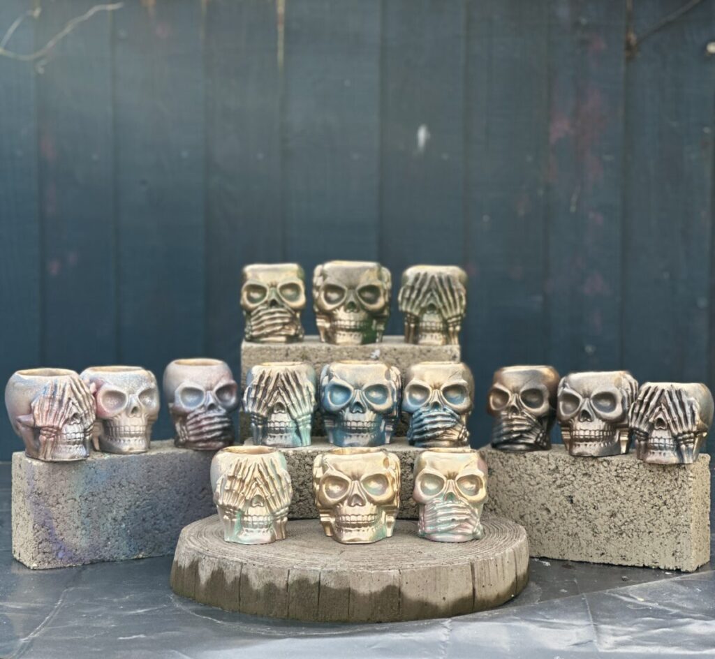A collection of small handcrafted skull planters in "See, Hear, and Speak No Evil" poses, featuring various iridescent and metallic finishes, displayed on concrete blocks and a wooden stump.