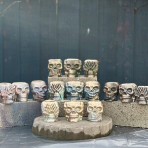 A collection of small handcrafted skull planters in "See, Hear, and Speak No Evil" poses, featuring various iridescent and metallic finishes, displayed on concrete blocks and a wooden stump.