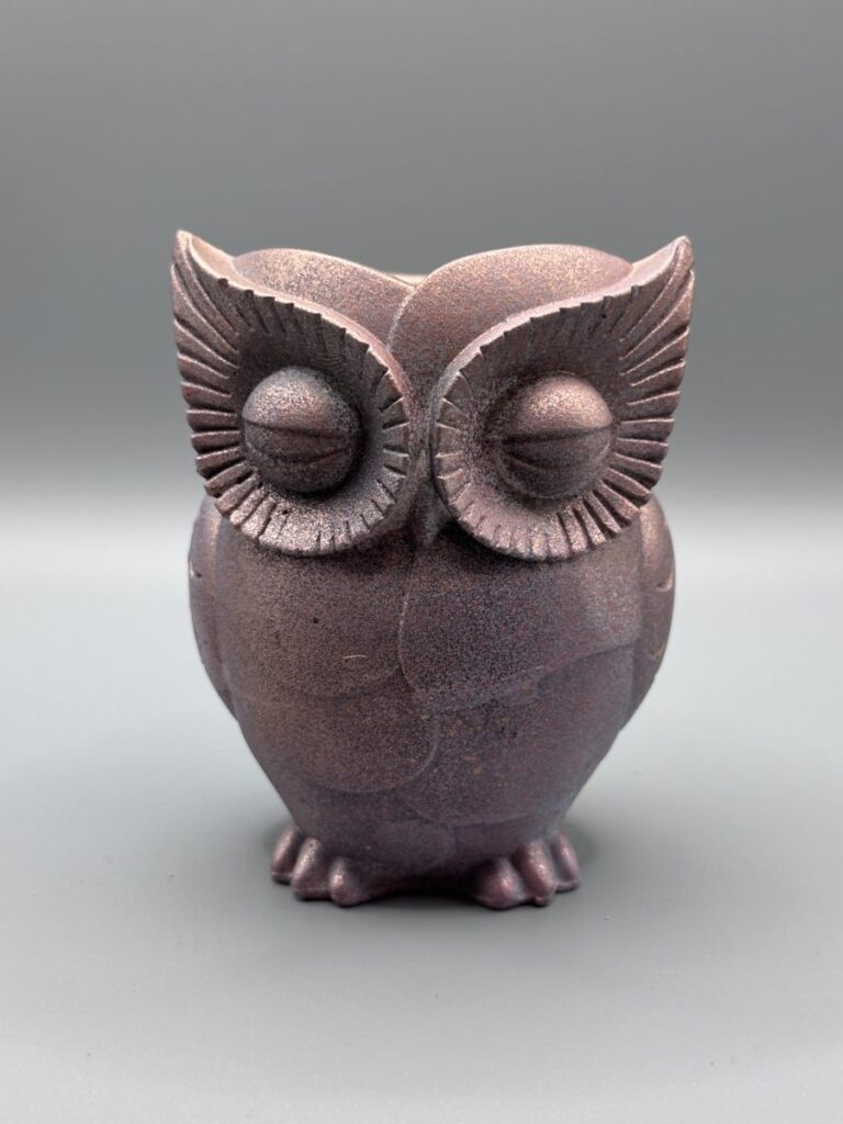 A small, hand-finished owl-shaped planter with a shimmering rose gold stone texture, resting on a rustic wood tree stump.