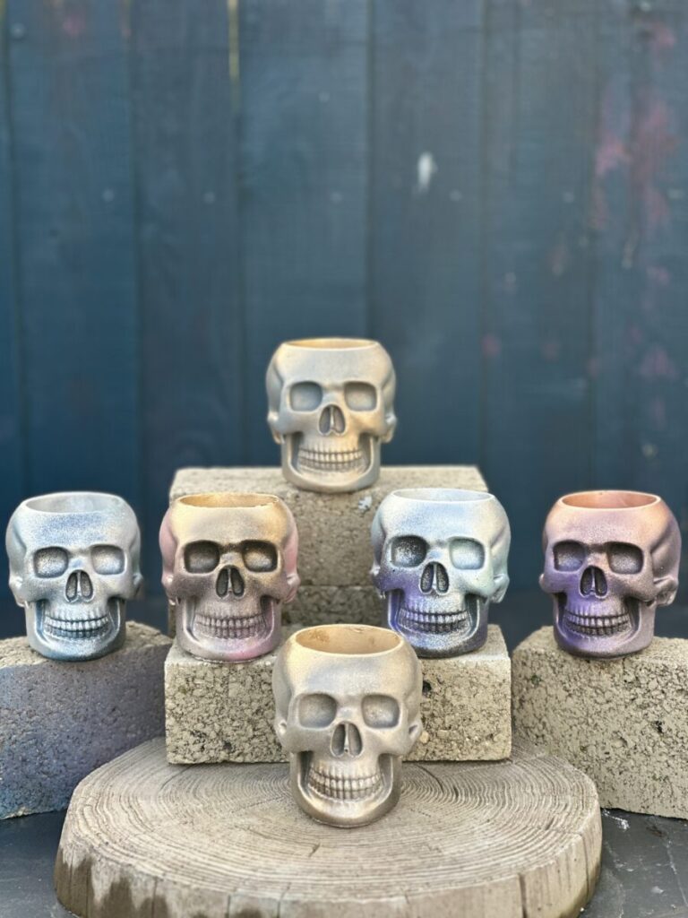 A collection of skull-shaped planters in various iridescent, gold, and silver finishes arranged on stone blocks.