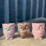 A handcrafted owl planter with a soft metallic rose-pink finish and intricate feather textures, displayed outdoors on a wooden tree stump.