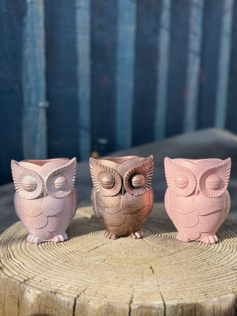 A handcrafted owl planter with a soft metallic rose-pink finish and intricate feather textures, displayed outdoors on a wooden tree stump.