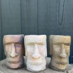 hree handmade cement Moai head planters in copper, pale sandstone and gold finishes on a concrete tree slice against a dark green wooden wall
