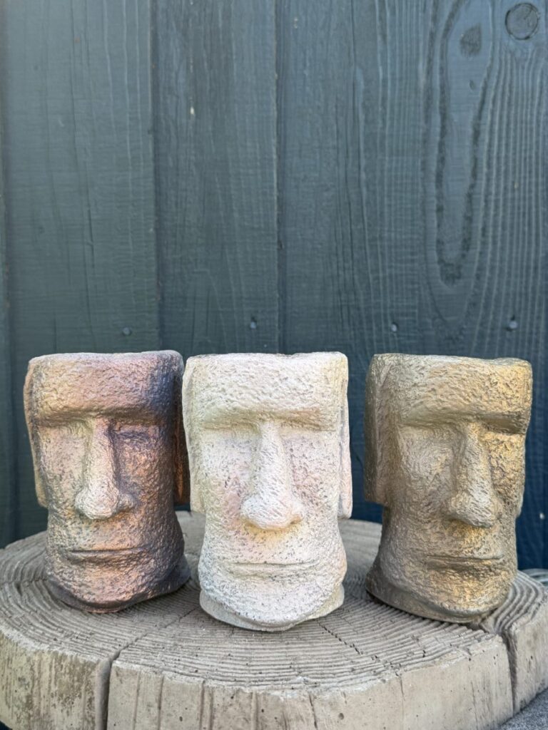 hree handmade cement Moai head planters in copper, pale sandstone and gold finishes on a concrete tree slice against a dark green wooden wall