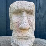 Handmade sandstone Moai head planter with speckled cement texture, blush pink cheeks and soft green shading, sitting on a concrete tree slice against a dark wooden wall.