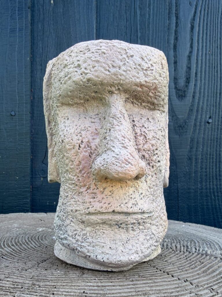 Handmade sandstone Moai head planter with speckled cement texture, blush pink cheeks and soft green shading, sitting on a concrete tree slice against a dark wooden wall.