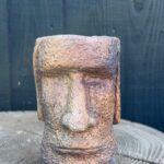 Handmade copper patina Moai head planter with rough cement texture, smoky plum shadows and gold highlights, sitting on a concrete tree slice against a dark wooden wall.