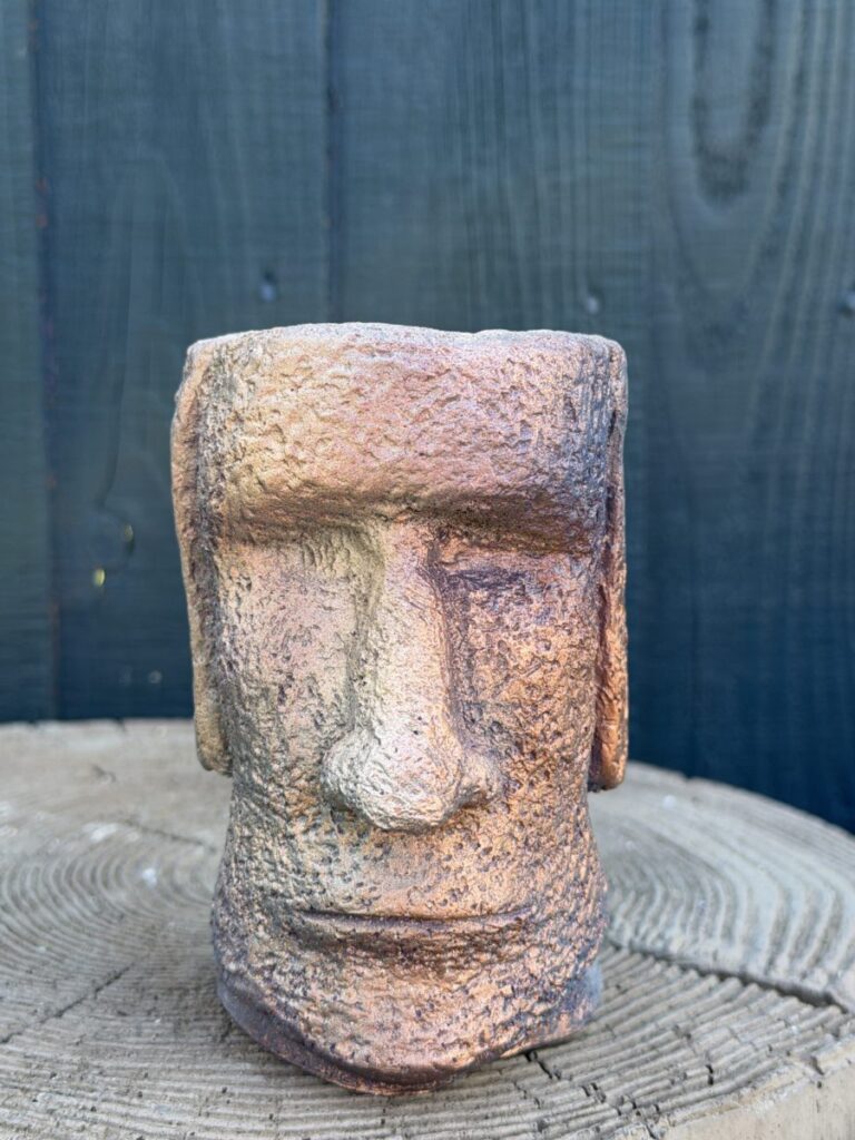 Handmade copper patina Moai head planter with rough cement texture, smoky plum shadows and gold highlights, sitting on a concrete tree slice against a dark wooden wall.