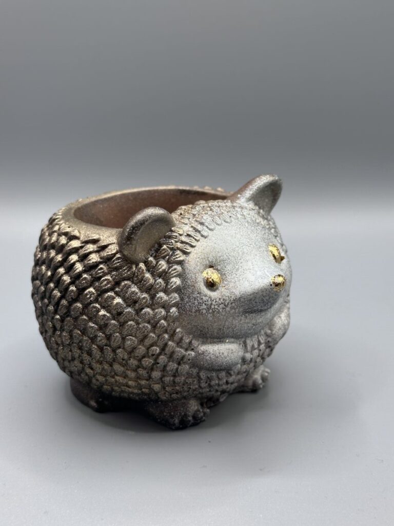A small, round hedgehog-shaped planter with a textured silver metallic finish and gold leaf accents on its eyes and nose, centered on a grey background.