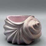 A spiral-shaped ceramic shell vessel with a shimmering iridescent champagne and soft pink finish, showcasing detailed ridges and a textured surface against a neutral background.