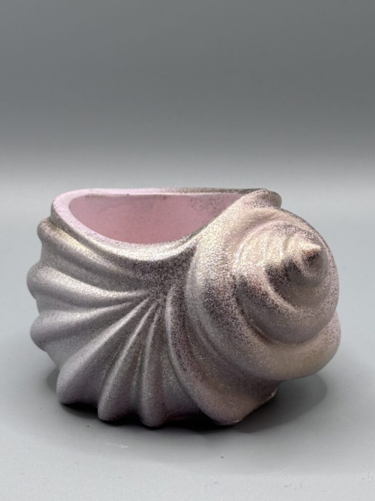 A spiral-shaped ceramic shell vessel with a shimmering iridescent champagne and soft pink finish, showcasing detailed ridges and a textured surface against a neutral background.
