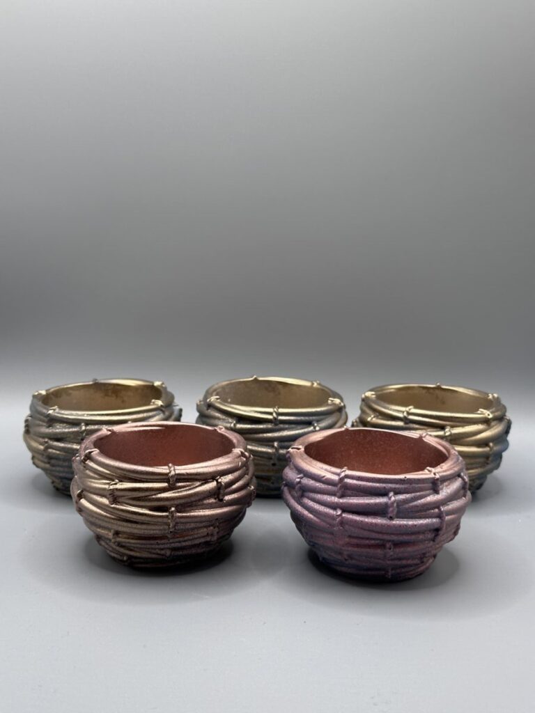 A group of five small, round planters with a heavy woven texture, displayed in a variety of metallic gradient colors including purple, copper, gold, and blue-bronze.