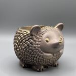 A small, round hedgehog planter with a warm metallic bronze-brown finish, detailed textured quills, and bright gold-leaf accents on the eyes and nose.