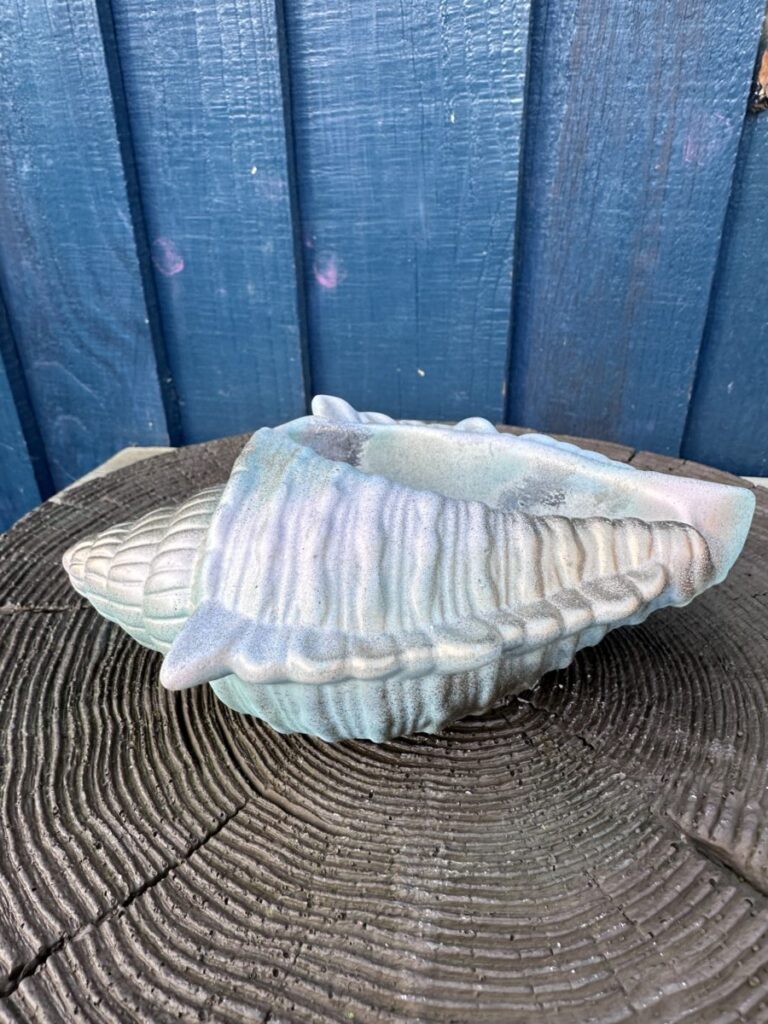 An iridescent hand-cast planter shaped like a large conch shell with a seafoam blue and pink pearlescent finish, resting on a dark wood grain surface against a blue wooden wall.