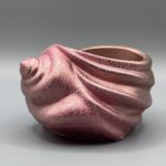 A spiral seashell-shaped ceramic vessel with a shimmering rose gold and pink metallic finish, featuring textured ridges and a deep interior, set against a grey background.