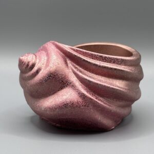 A spiral seashell-shaped ceramic vessel with a shimmering rose gold and pink metallic finish, featuring textured ridges and a deep interior, set against a grey background.