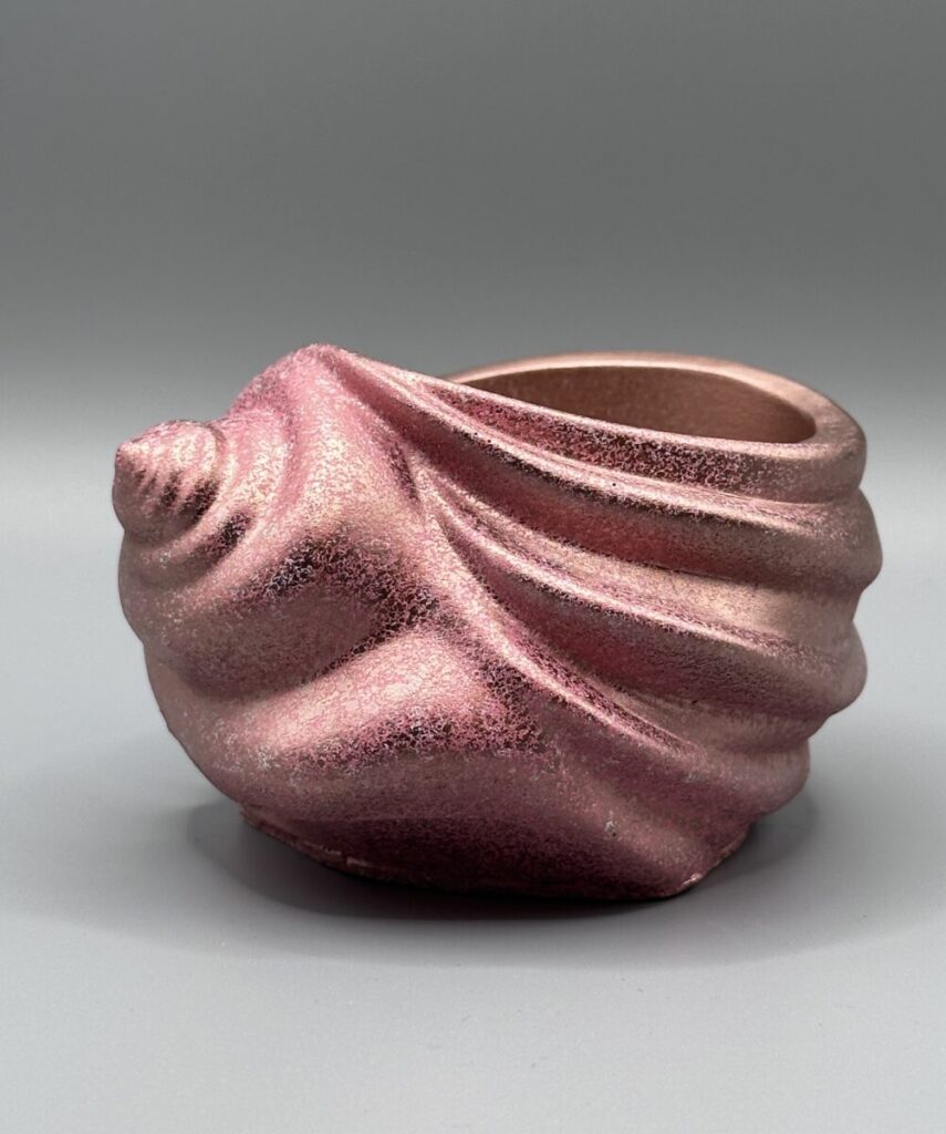 A spiral seashell-shaped ceramic vessel with a shimmering rose gold and pink metallic finish, featuring textured ridges and a deep interior, set against a grey background.
