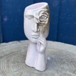 A surrealist face-shaped planter in blush pink with a textured sandstone finish, featuring a sculpted hand holding a gold-leaf rose over one eye, set against a blue wooden background.
