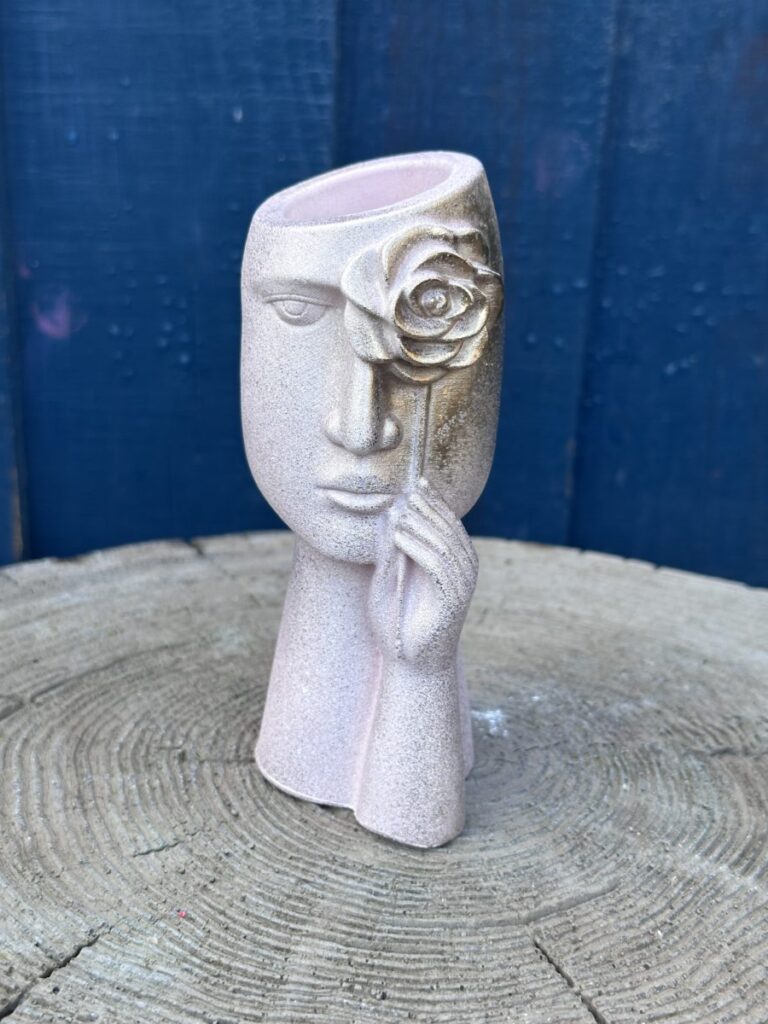 A surrealist face-shaped planter in blush pink with a textured sandstone finish, featuring a sculpted hand holding a gold-leaf rose over one eye, set against a blue wooden background.