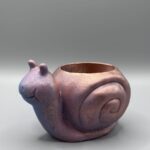 A small, snail-shaped succulent planter with an iridescent metallic finish shifting from purple to copper, featuring a stippled texture and a smiling face against a gray background.