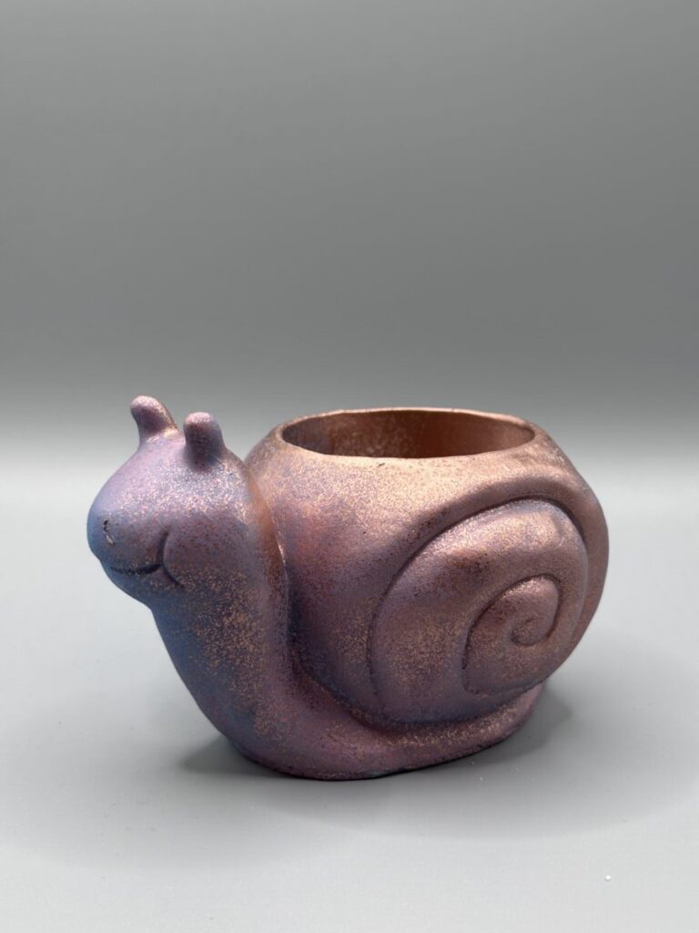 A small, snail-shaped succulent planter with an iridescent metallic finish shifting from purple to copper, featuring a stippled texture and a smiling face against a gray background.