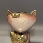 A cute fox-shaped planter with a crown and a large leaf finished in metallic gold, featuring a terracotta and cream textured surface, displayed against a gray background.