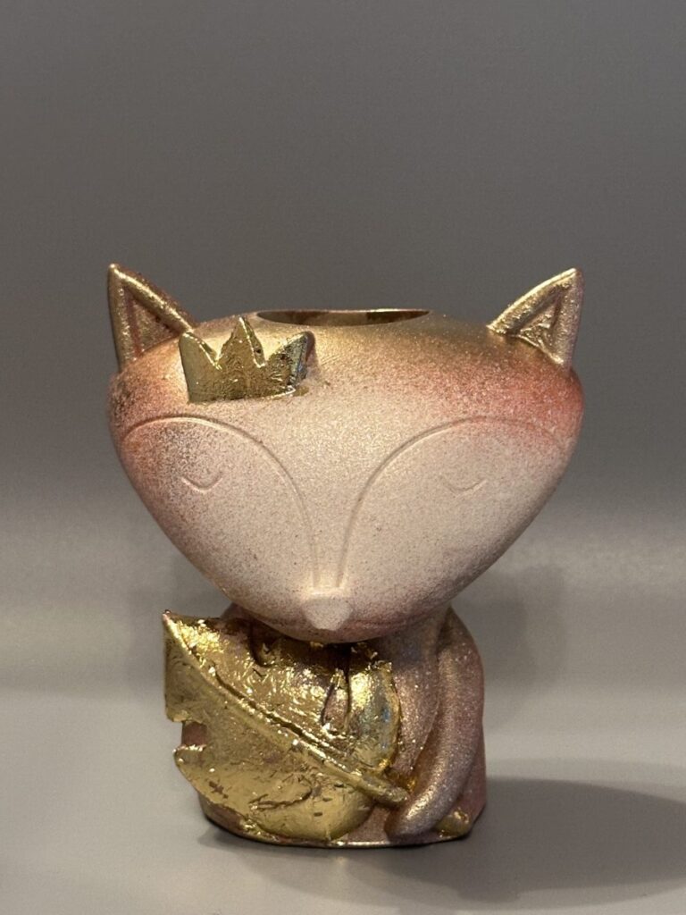 A cute fox-shaped planter with a crown and a large leaf finished in metallic gold, featuring a terracotta and cream textured surface, displayed against a gray background.