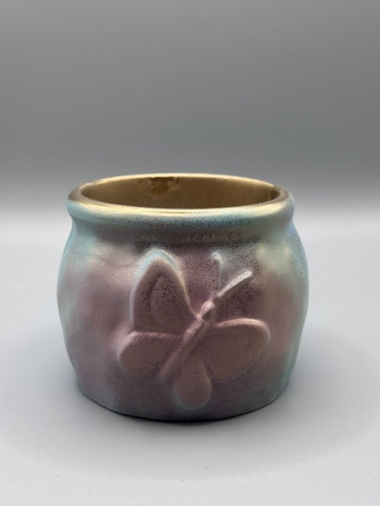 A small, rounded ceramic-style pot with an embossed butterfly on the side, featuring a pink and blue watercolor finish and a metallic gold rim.
