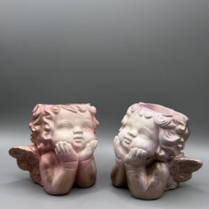 Two small cherub-shaped planters with detailed wings and curly hair, finished in a pink and metallic gold ombré, resting on their hands against a grey background.