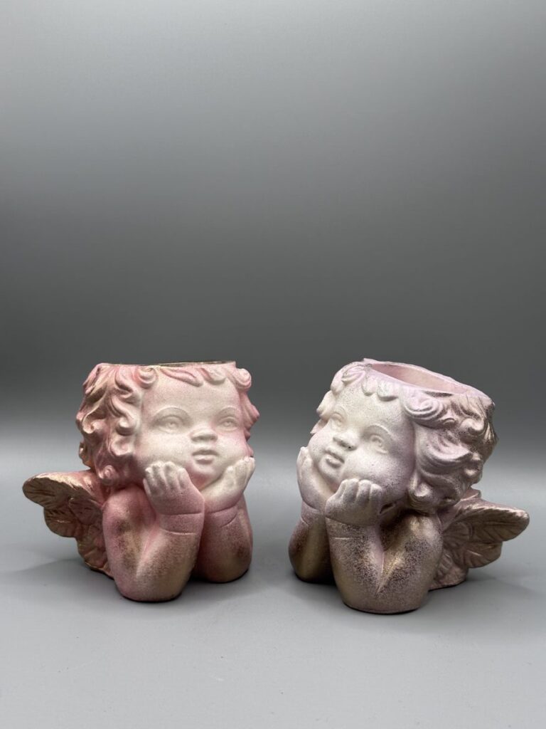 Two small cherub-shaped planters with detailed wings and curly hair, finished in a pink and metallic gold ombré, resting on their hands against a grey background.