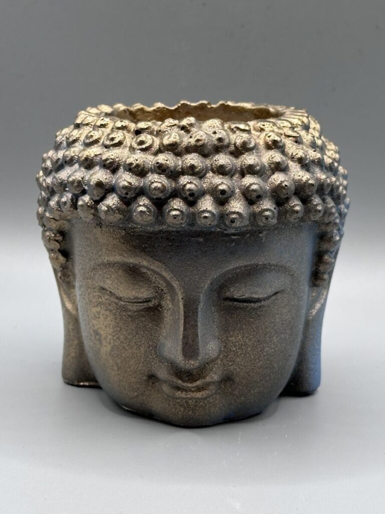 A detailed planter shaped like a Buddha head with a weathered bronze and gold finish, showing eyes closed in a meditative expression against a grey background.