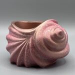 A spiral seashell-shaped ceramic vessel with a shimmering rose gold and pink metallic finish, featuring textured ridges and a deep interior, set against a grey background.