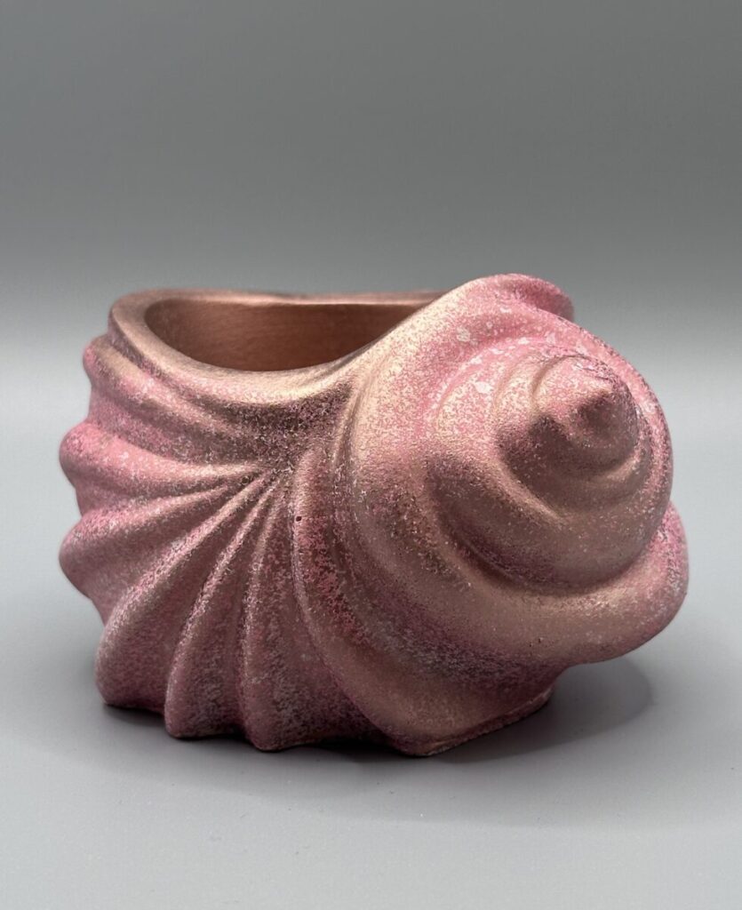 A spiral seashell-shaped ceramic vessel with a shimmering rose gold and pink metallic finish, featuring textured ridges and a deep interior, set against a grey background.
