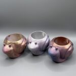Three small, smiling snail-shaped plant pots in various iridescent metallic finishes (gold-pink, purple-silver, and blue-pink) arranged on a grey background.