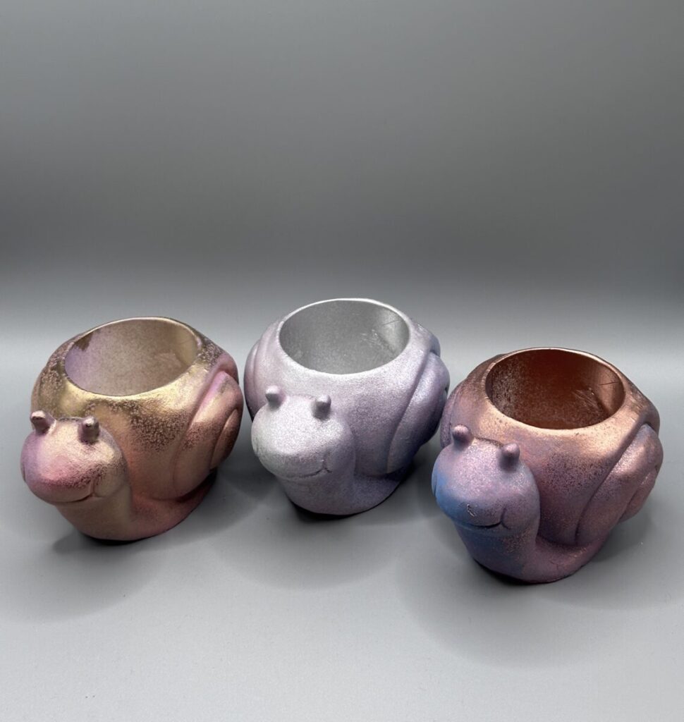 Three small, smiling snail-shaped plant pots in various iridescent metallic finishes (gold-pink, purple-silver, and blue-pink) arranged on a grey background.