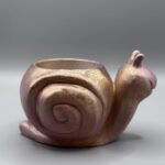 A small, smiling snail-shaped plant pot with a shimmering rose-gold and amber metallic finish against a grey background