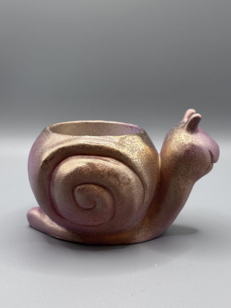 A small, smiling snail-shaped plant pot with a shimmering rose-gold and amber metallic finish against a grey background