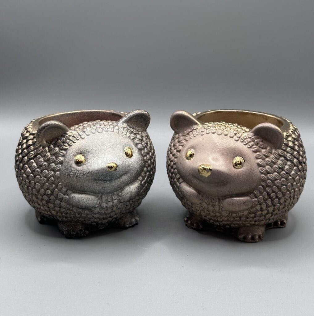 Two small, round hedgehog planters side-by-side; one is a metallic silver and the other is a metallic rose-bronze, both featuring textured quills and gold-leaf eyes and noses.
