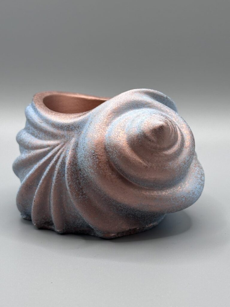 A spiral conch-shaped ceramic vessel with a dual-toned metallic finish featuring warm copper highlights and cool slate-blue undertones, set against a grey background.