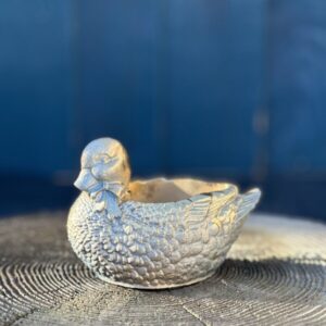 A small planter shaped like a mallard duck with a bow-tie, finished in metallic gold and resting on a weathered wooden tree stump against a blue background.