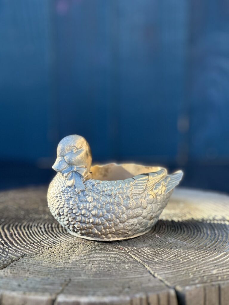 A small planter shaped like a mallard duck with a bow-tie, finished in metallic gold and resting on a weathered wooden tree stump against a blue background.