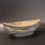 A boat-shaped ceramic vessel with a metallic golden-bronze glaze and raised rope textures, shown in two different lighting setups to highlight the shimmering finish.