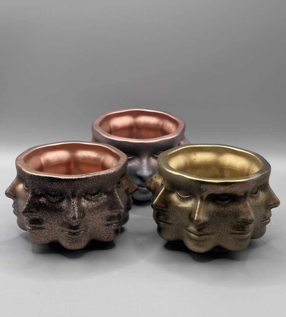 Three circular plant pots featuring multiple repeating faces sculpted around the exterior, shown in bronze, copper, and iridescent purple metallic finishes