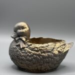 A small planter shaped like a mallard duck with a bow-tie, finished in metallic gold and resting on a gray background.