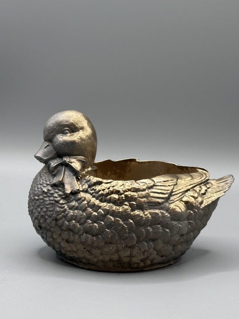 A small planter shaped like a mallard duck with a bow-tie, finished in metallic gold and resting on a gray background.
