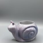 A small, smiling snail-shaped plant pot with a shimmering iridescent lavender and light blue metallic finish against a grey background.